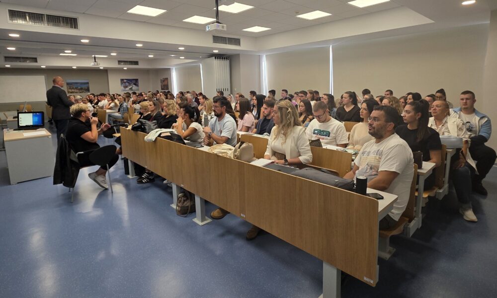 Na fotografiji je prikazana radionica ili predavanje u fakultetskoj učionici. U prostranoj i moderno opremljenoj dvorani nalazi se veći broj studenata i drugih sudionika koji prate izlaganje. U prednjem dijelu prostorije vidi se predavač koji se obraća publici, dok je uz njega osoba koja sudjeluje u izlaganju. Učionica je opremljena projektorom, računalom i velikim ekranom. Sudionici sjede u nekoliko redova i pažljivo prate predavanje, a neki vode bilješke. Fotografija dobro prikazuje aktivno sudjelovanje i zainteresiranost sudionika te atmosferu akademskog okupljanja i edukacije.