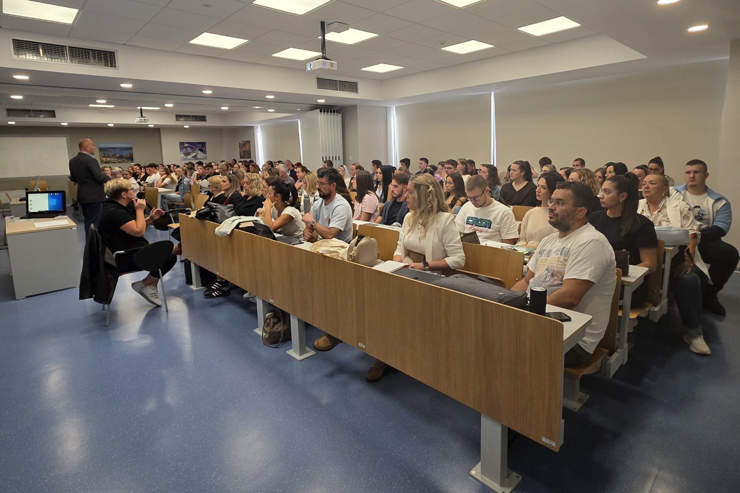 Na fotografiji je prikazana radionica ili predavanje u fakultetskoj učionici. U prostranoj i moderno opremljenoj dvorani nalazi se veći broj studenata i drugih sudionika koji prate izlaganje. U prednjem dijelu prostorije vidi se predavač koji se obraća publici, dok je uz njega osoba koja sudjeluje u izlaganju. Učionica je opremljena projektorom, računalom i velikim ekranom. Sudionici sjede u nekoliko redova i pažljivo prate predavanje, a neki vode bilješke. Fotografija dobro prikazuje aktivno sudjelovanje i zainteresiranost sudionika te atmosferu akademskog okupljanja i edukacije.