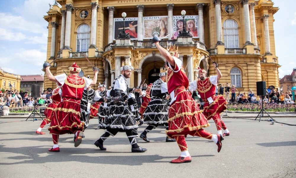 Folklorni plesači u raskošnim crvenim i crnim kostimima nastupaju ispred Hrvatskog narodnog kazališta u Zagrebu.
