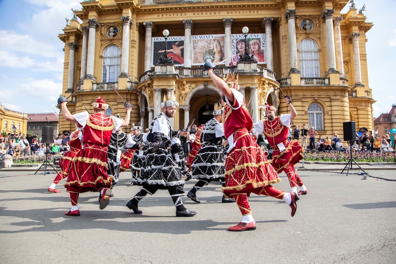 Folklorni plesači u raskošnim crvenim i crnim kostimima nastupaju ispred Hrvatskog narodnog kazališta u Zagrebu.