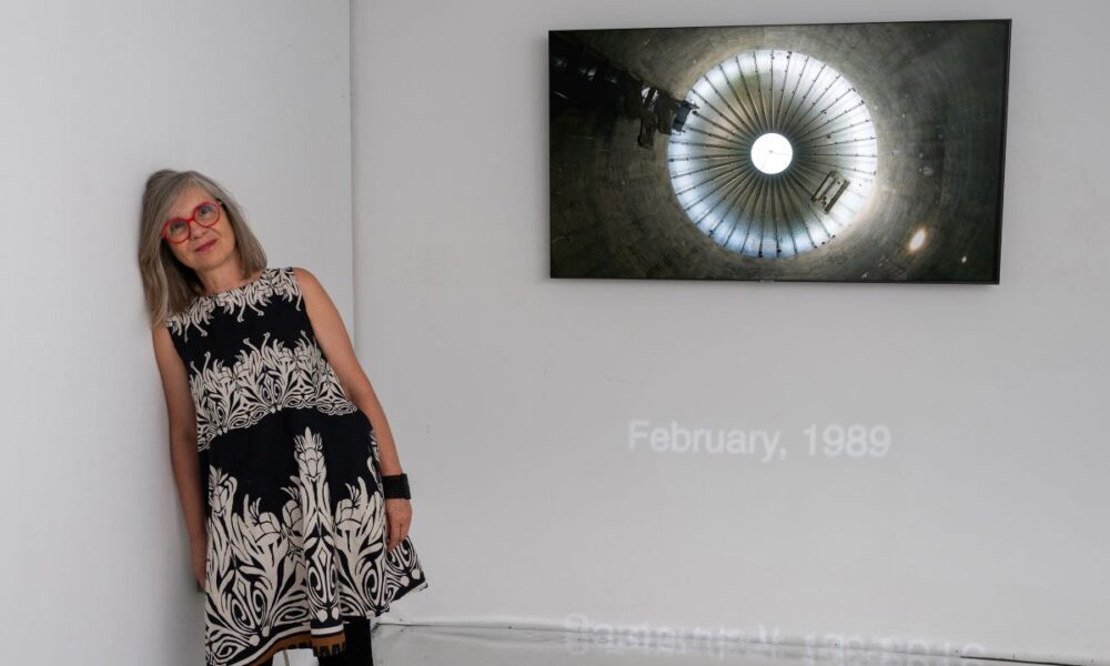 Žena s crvenim naočalama stoji uz zid galerije na kojem je izložena fotografija kružne arhitektonske strukture.