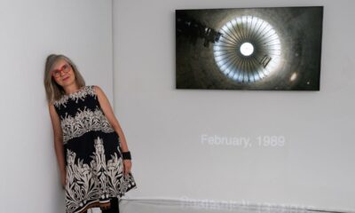 Žena s crvenim naočalama stoji uz zid galerije na kojem je izložena fotografija kružne arhitektonske strukture.