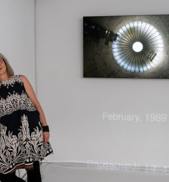 Žena s crvenim naočalama stoji uz zid galerije na kojem je izložena fotografija kružne arhitektonske strukture.