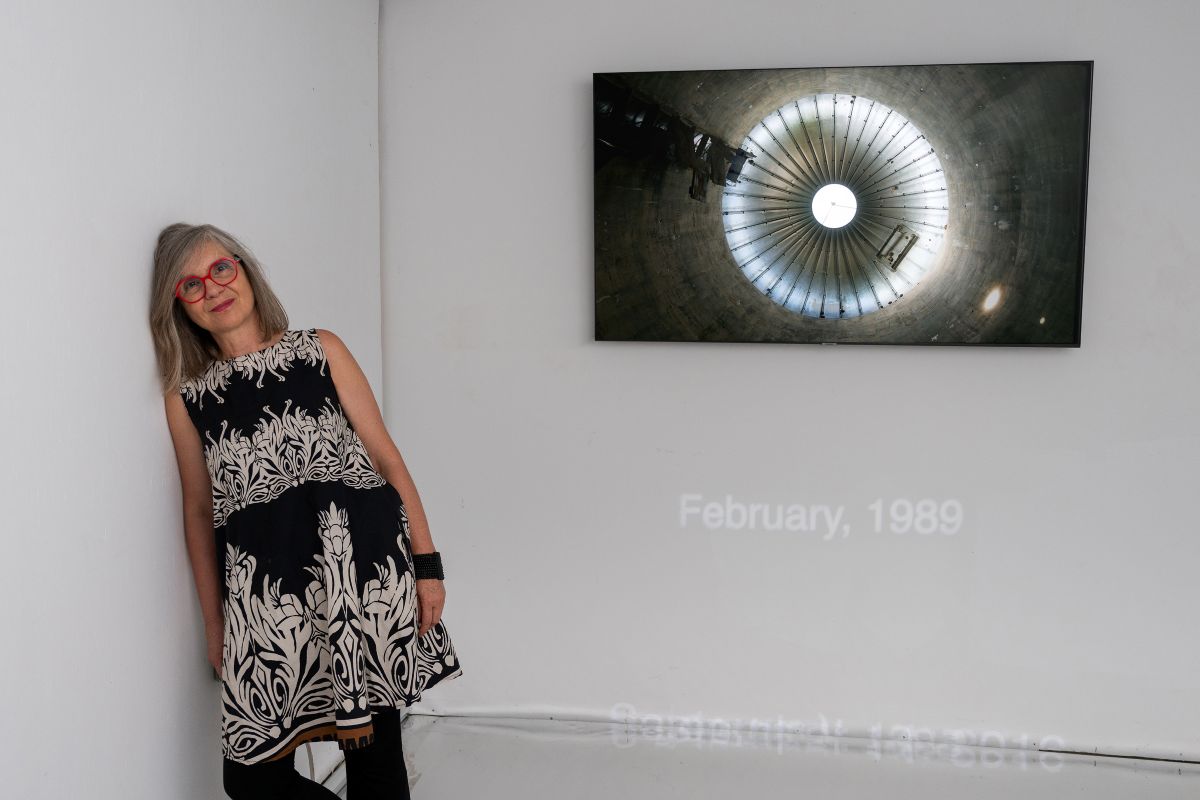 Žena s crvenim naočalama stoji uz zid galerije na kojem je izložena fotografija kružne arhitektonske strukture.