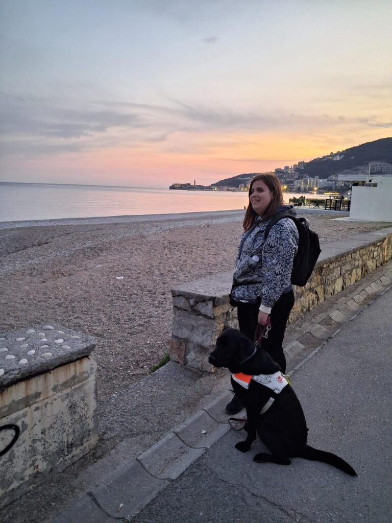 Žena s crnim psom pomagačem stoji uz šetnicu pokraj plaže u suton.