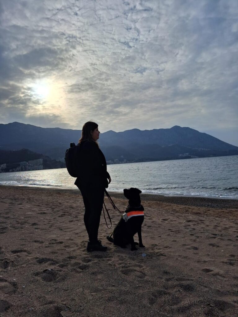 Žena i crni pas pomagač stoje na pješčanoj plaži, okrenuti prema moru i planinama.