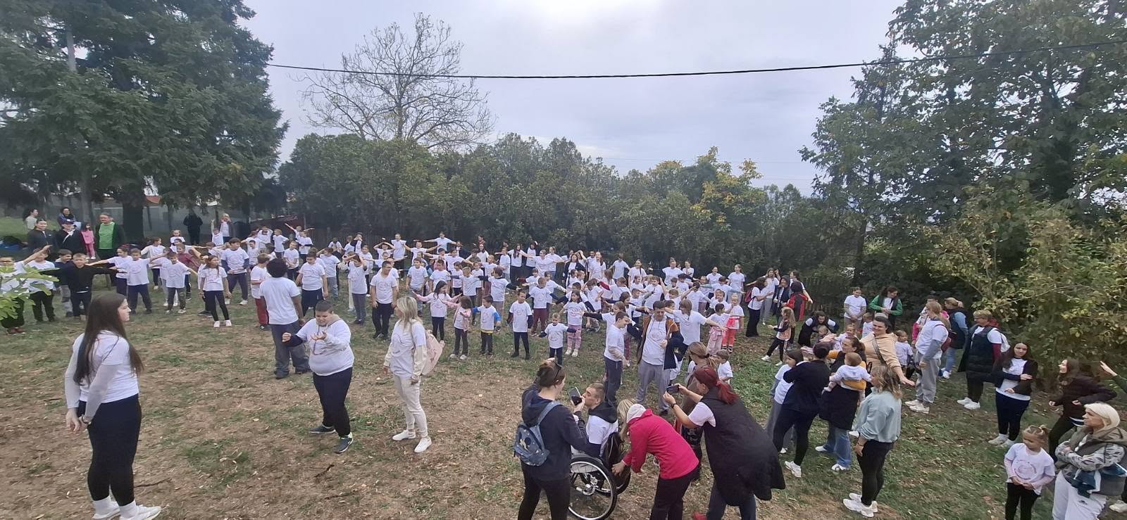 Na fotografiji se vidi velik broj djece, mladih i odraslih okupljenih na otvorenom, u zelenom okruženju okruženom drvećem. Sudionici nose bijele majice i sudjeluju u zajedničkim aktivnostima, dok volonteri i odrasli pomažu u organizaciji druženja. U prvom planu vidi se i osoba u invalidskim kolicima, što dodatno naglašava važnost uključivanja svih sudionika. Fotografija prikazuje zajedništvo, druženje i prihvaćanje različitosti, što su glavne vrijednosti akcije „Različiti, a jednaki“. Kroz zajedničke aktivnosti djeca i mladi uče o međusobnom poštovanju, solidarnosti i jednakim mogućnostima za sve.