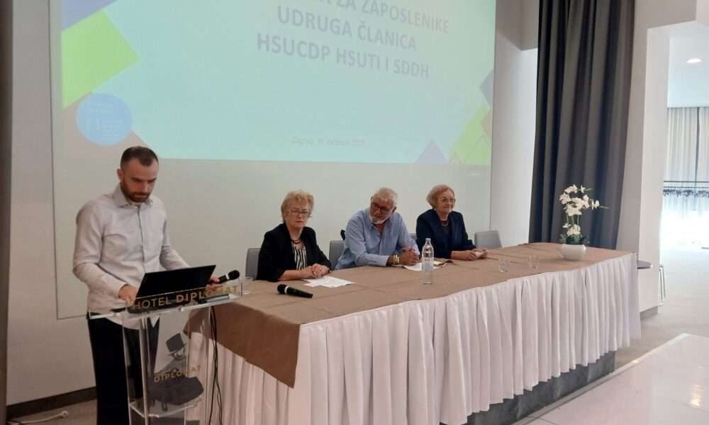 Sudionici radionice za zaposlenike udruga osoba s invaliditetom sjede za stolom, dok muškarac stoji za govornicom.