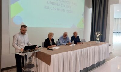 Sudionici radionice za zaposlenike udruga osoba s invaliditetom sjede za stolom, dok muškarac stoji za govornicom.
