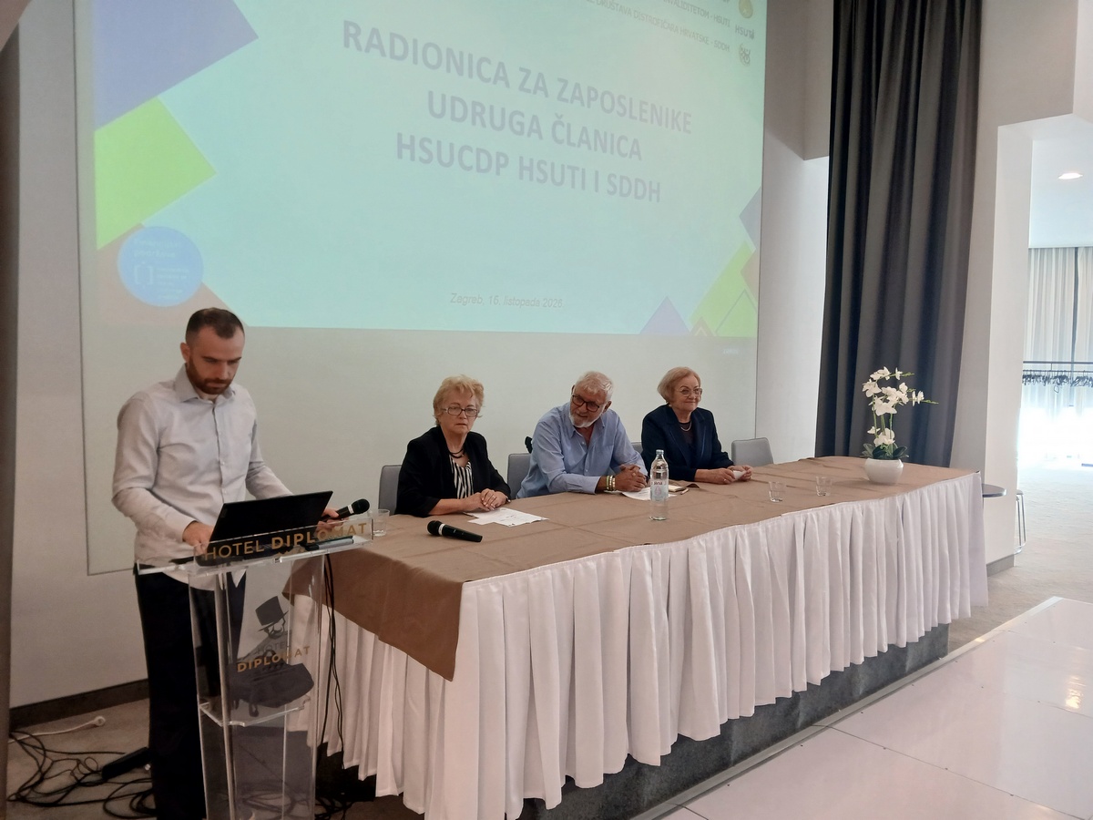 Sudionici radionice za zaposlenike udruga osoba s invaliditetom sjede za stolom, dok muškarac stoji za govornicom.