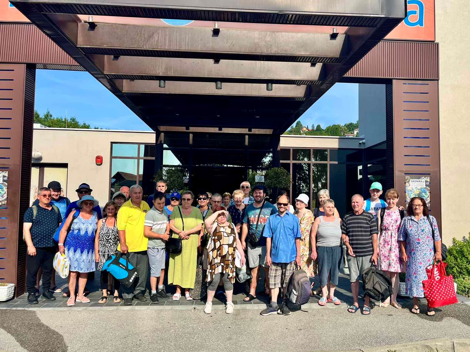 Na fotografiji je skupina članova Udruge slijepih Međimurske županije okupljena ispred ulaza u jedan objekt. Članovi su nasmijani i opušteni, a dio njih nosi šešire i sunčane naočale. Fotografija bilježi zajedničko druženje i izlet u vedroj, ljetnoj atmosferi.