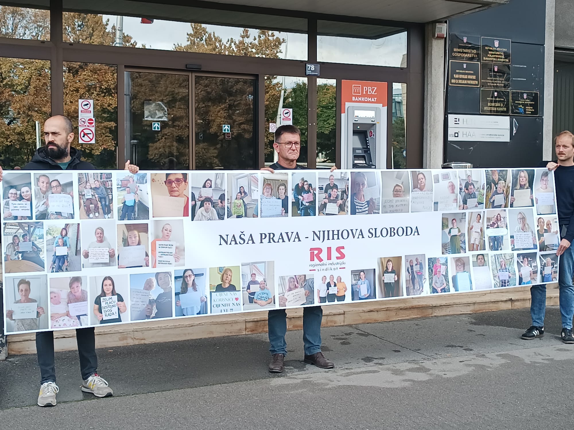 Na fotografiji su tri osobe koje ispred zgrade Ministarstva rada drže veliki transparent Regionalnog industrijskog sindikata (RIS). Na transparentu se nalazi velik broj fotografija osobnih asistenata i njihovih poruka, a u središnjem dijelu istaknuta je poruka „Naša prava – njihova sloboda“ uz logo RIS-a. Fotografije i poruke na transparentu prikazuju osobne asistente koji žele ukazati na svoj položaj i probleme s kojima se susreću u radu. U pozadini se vidi ulaz u zgradu Ministarstva, kao i bankomat te oznake institucija smještenih u zgradi. Fotografija tako vizualno prikazuje sindikalno okupljanje i poruku osobnih asistenata da njihov rad i prava moraju biti prepoznati i poštovani.