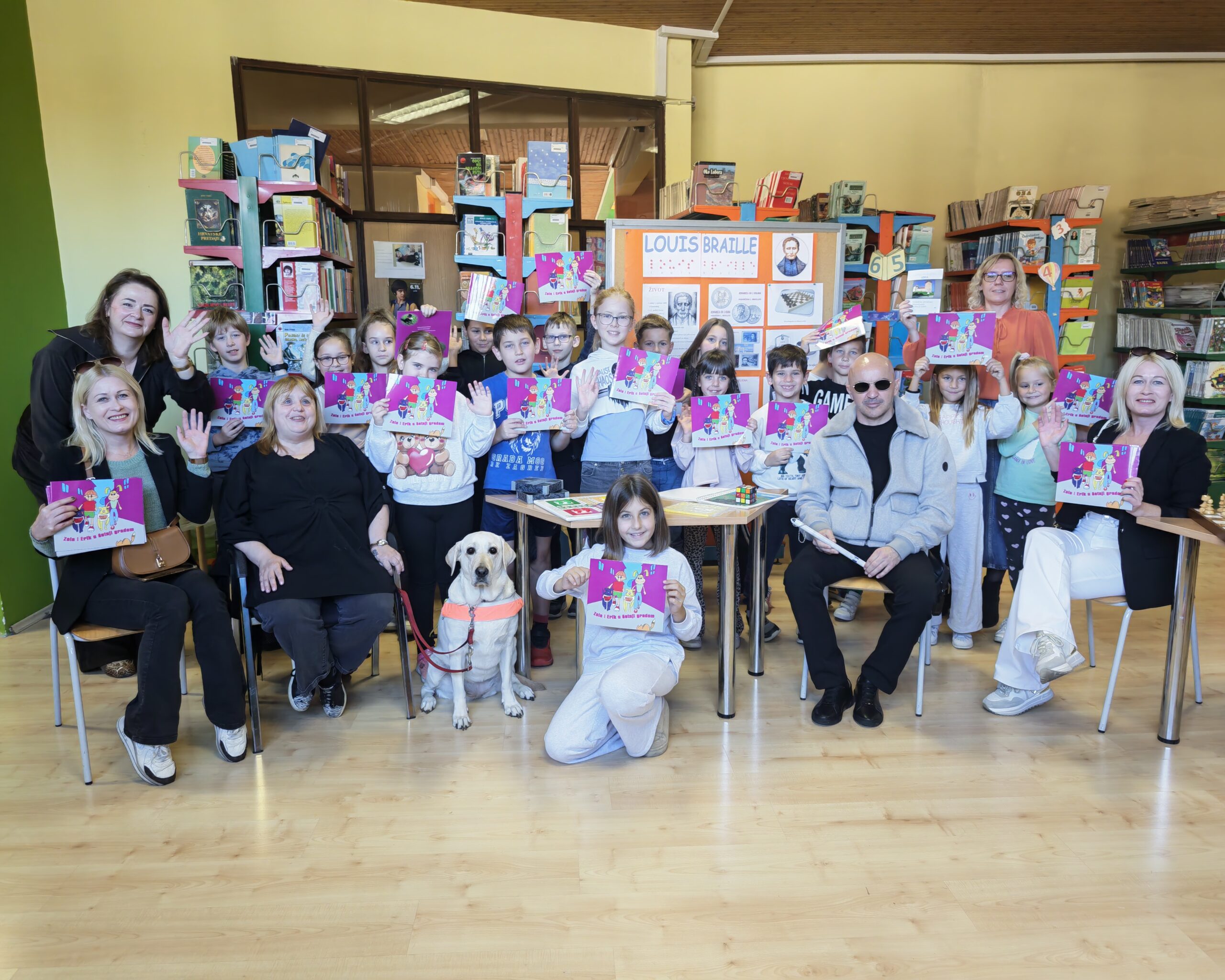 Na fotografiji su predstavnici Udruge slijepih USKA i učenici Osnovne škole Dubovac tijekom edukativnog susreta u knjižnici. Sudionici drže edukativne slikovnice „Zola i Erik u šetnji gradom“, a u prvom planu nalazi se pas vodič. Uz predstavljanje Brailleova pisma, bijelog štapa i pomagala za slijepe osobe, djeca su imala priliku upoznati se sa svakodnevnim životom osoba s oštećenjem vida. Susret je protekao u veselom i prijateljskom ozračju, s ciljem podizanja svijesti o važnosti razumijevanja, pristupačnosti i uključivanja slijepih i slabovidnih osoba u društvo.