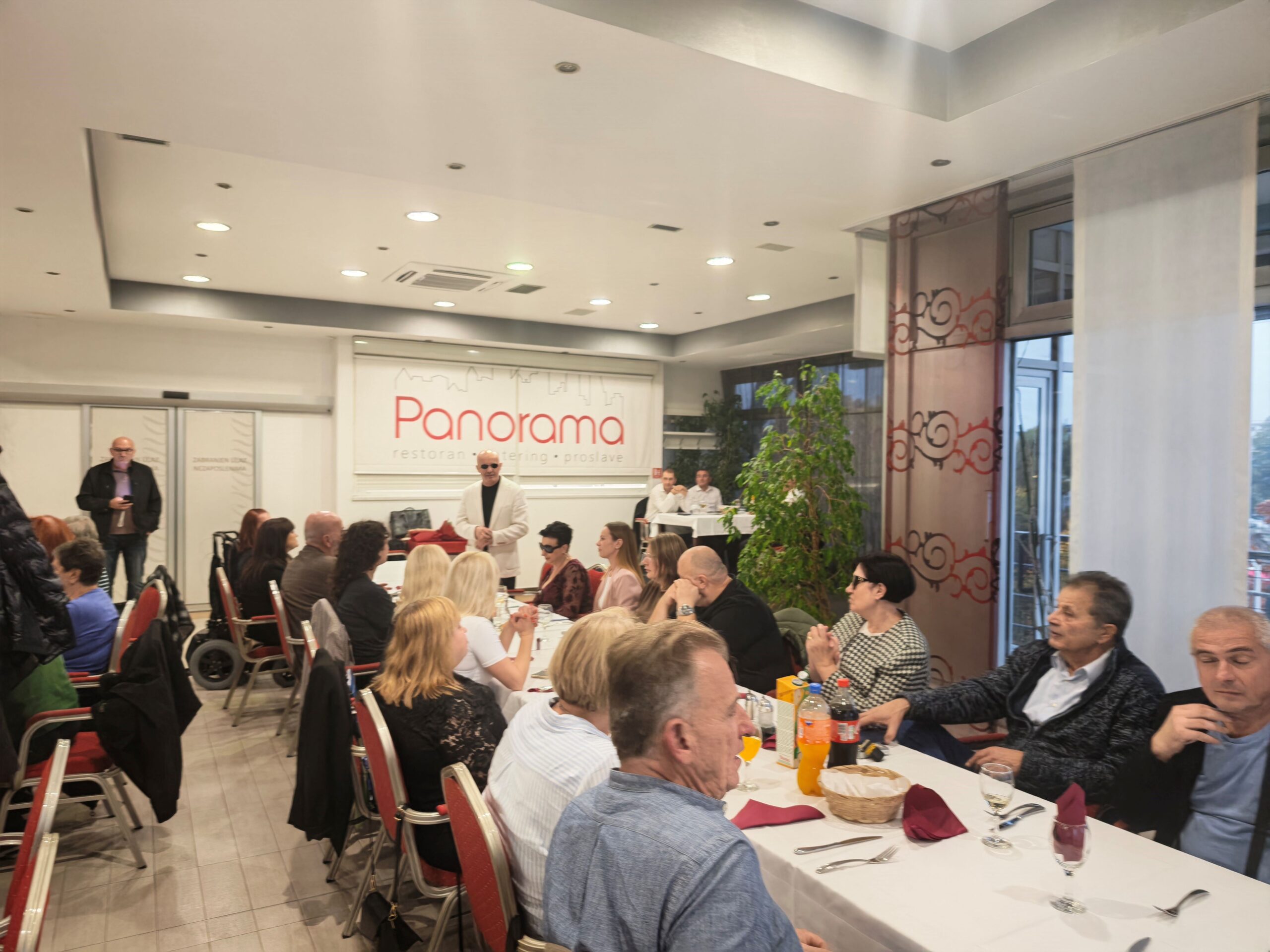 Fotografija prikazuje članove i goste Udruge slijepih USKA tijekom godišnje skupštine i zajedničkog druženja u Restoranu Panorama u Karlovcu. Sudionici sjede za svečano postavljenim stolovima i prate program, a u pozadini se vidi predsjednik udruge. Ugodna atmosfera i okupljanje za istim stolom ističu važnost zajedništva, druženja i međusobne podrške.
