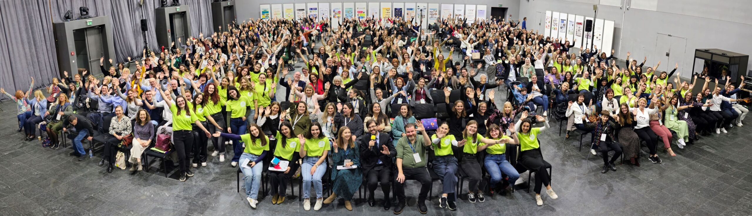 Na fotografiji je velika skupina sudionika ATAAC konferencije, okupljena u konferencijskoj dvorani. Gotovo svi sudionici veselo poziraju prema kameri s podignutim rukama, stvarajući vrlo energičnu i pozitivnu atmosferu. U prvom planu su organizatori i članovi volonterskog tima u prepoznatljivim zelenim majicama, dok se u pozadini vide brojni sudionici, izlagački panoi i prostor konferencije. Fotografija dobro dočarava zajedništvo, velik odaziv i opuštenu atmosferu ATAAC-a.
