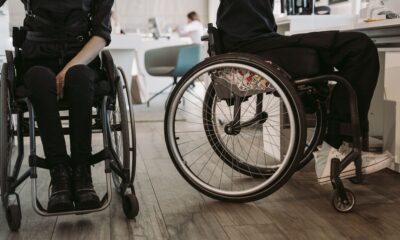 Na fotografiji su prikazane dvije osobe u invalidskim kolicima u zatvorenom prostoru koji nalikuje zdravstvenoj ustanovi ili rehabilitacijskom centru. Obje osobe odjevene su u tamnu odjeću, a u prvom planu posebno se ističu kotači invalidskih kolica. U pozadini se nalaze stolice, namještaj i druge osobe, što upućuje na prostor u kojem se odvijaju svakodnevne aktivnosti ili pruža određena vrsta podrške. Fotografija naglašava važnost pristupačnosti, mobilnosti i uključivanja osoba s invaliditetom u svakodnevni život. Može se povezati s temama poput prava osoba s invaliditetom, prilagođenog okruženja i ravnopravnog sudjelovanja u društvu.