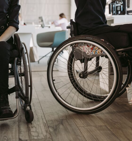 Na fotografiji su prikazane dvije osobe u invalidskim kolicima u zatvorenom prostoru koji nalikuje zdravstvenoj ustanovi ili rehabilitacijskom centru. Obje osobe odjevene su u tamnu odjeću, a u prvom planu posebno se ističu kotači invalidskih kolica. U pozadini se nalaze stolice, namještaj i druge osobe, što upućuje na prostor u kojem se odvijaju svakodnevne aktivnosti ili pruža određena vrsta podrške. Fotografija naglašava važnost pristupačnosti, mobilnosti i uključivanja osoba s invaliditetom u svakodnevni život. Može se povezati s temama poput prava osoba s invaliditetom, prilagođenog okruženja i ravnopravnog sudjelovanja u društvu.