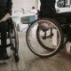Na fotografiji su prikazane dvije osobe u invalidskim kolicima u zatvorenom prostoru koji nalikuje zdravstvenoj ustanovi ili rehabilitacijskom centru. Obje osobe odjevene su u tamnu odjeću, a u prvom planu posebno se ističu kotači invalidskih kolica. U pozadini se nalaze stolice, namještaj i druge osobe, što upućuje na prostor u kojem se odvijaju svakodnevne aktivnosti ili pruža određena vrsta podrške. Fotografija naglašava važnost pristupačnosti, mobilnosti i uključivanja osoba s invaliditetom u svakodnevni život. Može se povezati s temama poput prava osoba s invaliditetom, prilagođenog okruženja i ravnopravnog sudjelovanja u društvu.