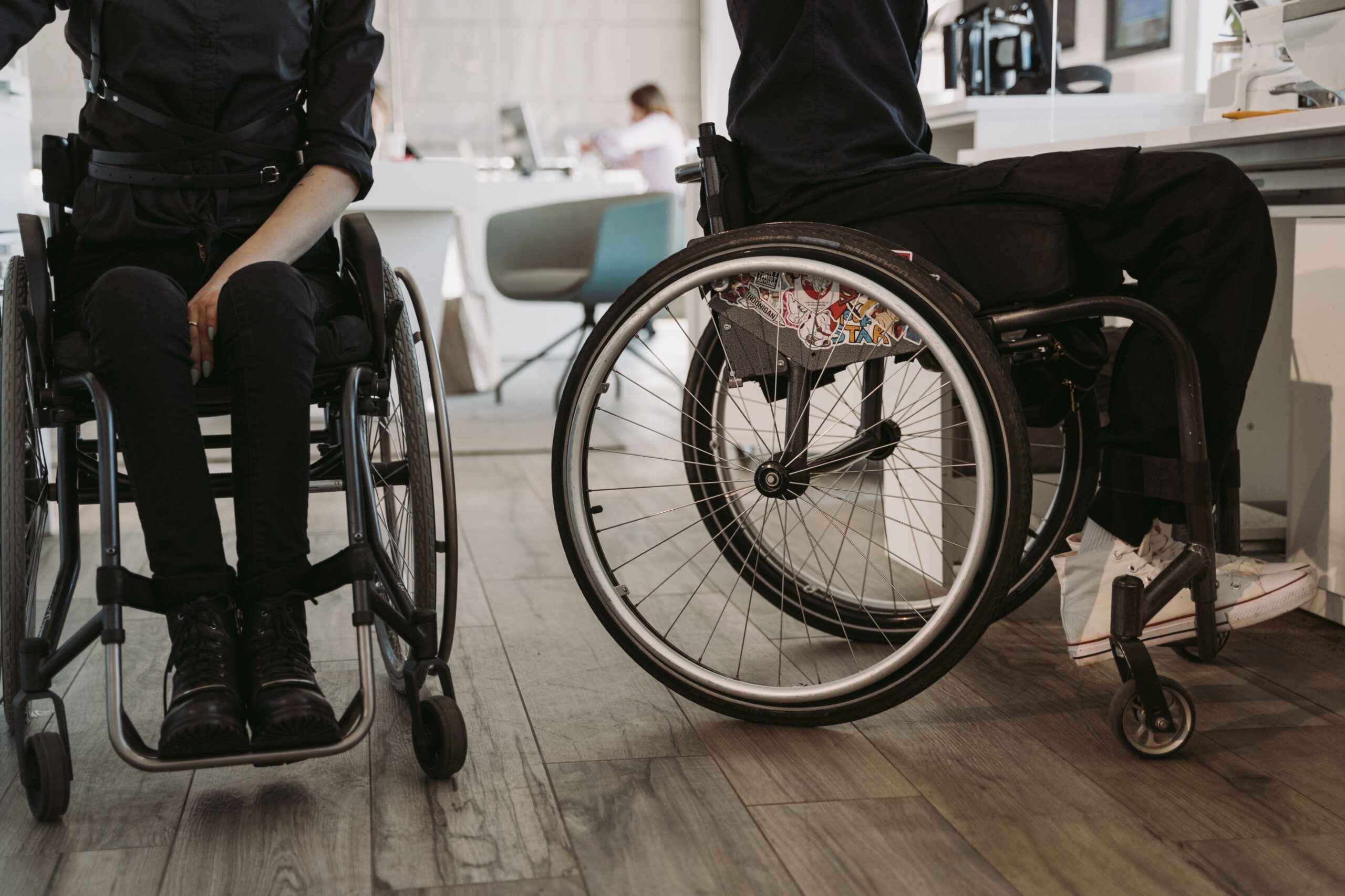 Na fotografiji su prikazane dvije osobe u invalidskim kolicima u zatvorenom prostoru koji nalikuje zdravstvenoj ustanovi ili rehabilitacijskom centru. Obje osobe odjevene su u tamnu odjeću, a u prvom planu posebno se ističu kotači invalidskih kolica. U pozadini se nalaze stolice, namještaj i druge osobe, što upućuje na prostor u kojem se odvijaju svakodnevne aktivnosti ili pruža određena vrsta podrške. Fotografija naglašava važnost pristupačnosti, mobilnosti i uključivanja osoba s invaliditetom u svakodnevni život. Može se povezati s temama poput prava osoba s invaliditetom, prilagođenog okruženja i ravnopravnog sudjelovanja u društvu.