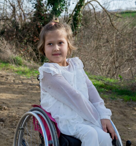 Na fotografiji je djevojčica koja sjedi u invalidskim kolicima na zemljanom putu u prirodi. Odjevena je u bijelu odjeću s nježnim uzorkom zvjezdica, a kosu nosi svezanu u rep. Na licu ima blag osmijeh i pogled usmjeren u stranu. U pozadini se nalaze grmlje, drveće i zelenilo, a sunčeva svjetlost stvara ugodan, prirodan ugođaj. Fotografija prikazuje djevojčicu u mirnom trenutku na otvorenom.