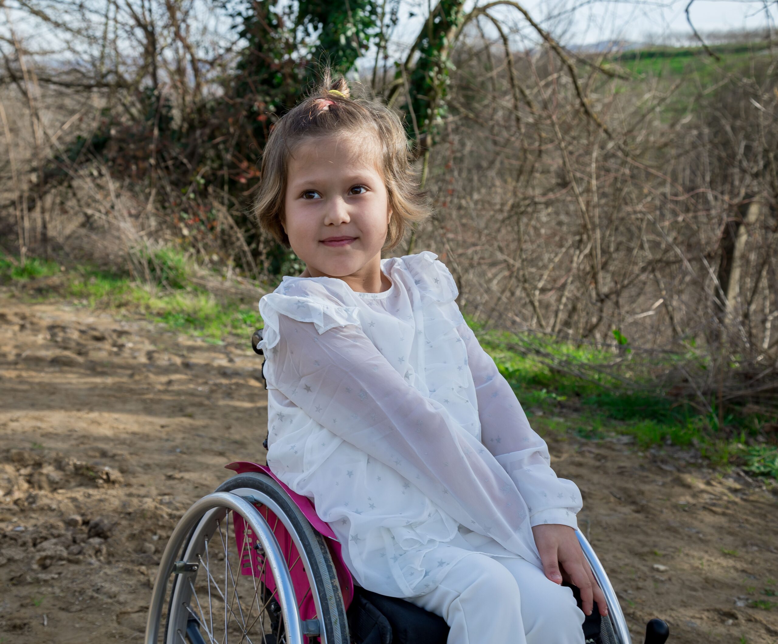 Na fotografiji je djevojčica koja sjedi u invalidskim kolicima na zemljanom putu u prirodi. Odjevena je u bijelu odjeću s nježnim uzorkom zvjezdica, a kosu nosi svezanu u rep. Na licu ima blag osmijeh i pogled usmjeren u stranu. U pozadini se nalaze grmlje, drveće i zelenilo, a sunčeva svjetlost stvara ugodan, prirodan ugođaj. Fotografija prikazuje djevojčicu u mirnom trenutku na otvorenom.